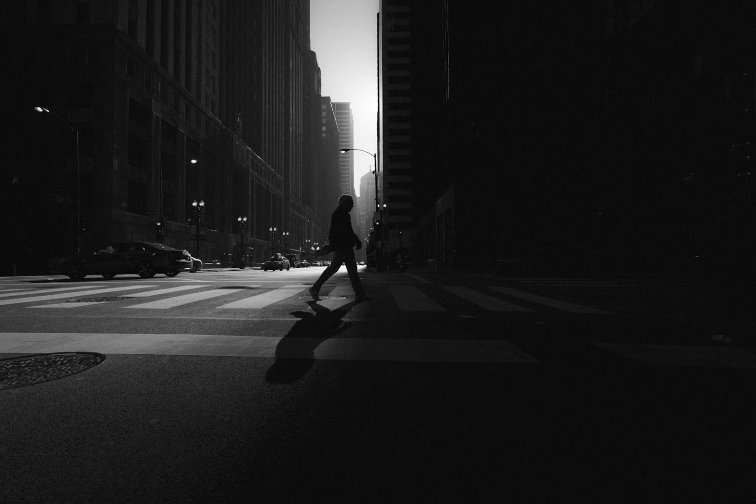 Study Finds EV Pedestrian Casualty Rate Comparable to Conventional Cars in UK Black and white silhouette of a person crossing a city street with tall buildings lining the roadway.