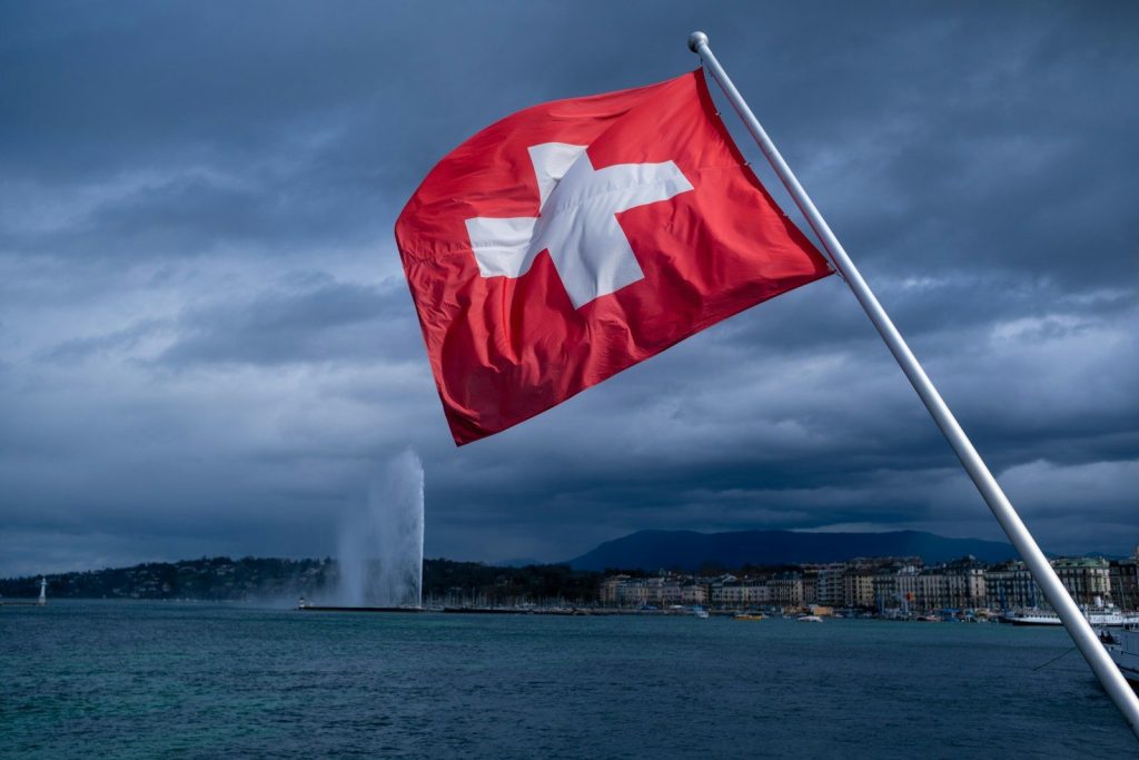 Swiss Canton’s New Tax Rules Leave Many Electric Car Owners Paying More Than Petrol Drivers a swiss flag blowing in the wind near a body of water